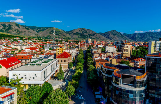 Vibrant Panoramic View of Kor&ccedil;&euml;, Albania with Colorful Rooftops, Tree-Lined Boulevards, and Majestic Mountain Range Under a Clear Blue Summer Sky