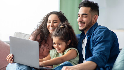 Young Arabic Parents And Their Little Girl Relaxing With Laptop At Home, Sitting On Couch In Living Room And Using Computer Together, Making Online Shopping Or Watching Family Movies, Closeup
