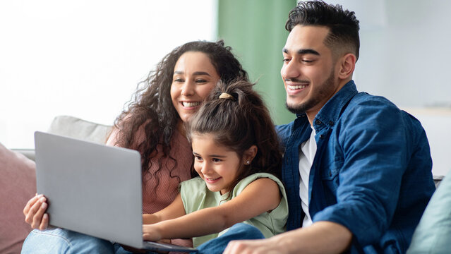 Young Arabic Parents And Their Little Girl Relaxing With Laptop At Home, Sitting On Couch In Living Room And Using Computer Together, Making Online Shopping Or Watching Family Movies, Closeup