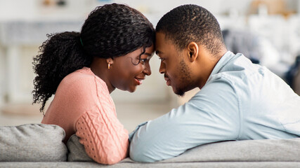 Loving black couple cuddling while sitting on couch at home, young african american man and woman in casual touching each other with foreheads and smiling, side view, closeup portrait