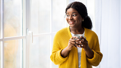Pretty black lady in casual standing next to window in living room, looking outside and drinking tea, copy space. Happy african american young woman enjoying sunny weather, drinking coffee
