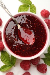 Sweet raspberry jam in bowl, berries and green leaves on white wooden table, flat lay