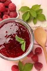 Sweet raspberry jam in glass jar, berries and mint on pink wooden table, flat lay