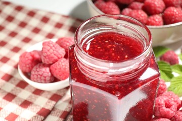 Delicious raspberry jam in glass jar, fresh berries, mint and checkered napkin on white table, closeup