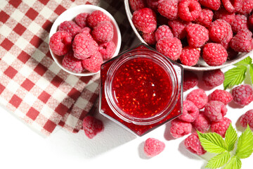 Delicious raspberry jam in glass jar, fresh berries, mint and checkered napkin on white table, flat lay