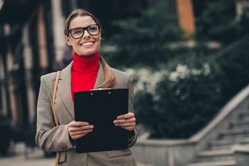 Confident businesswoman holding clipboard outdoors on a bright day in urban setting with modern buildings and greenery