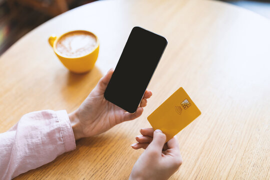 Contactless payment during coronavirus quarantine. Woman holding cellphone and credit card, drinking morning coffee in cafe interior, mockup on cellphone screen
