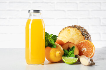Tasty juice in glass bottle, fresh fruits, ginger and mint on white table against brick wall, closeup