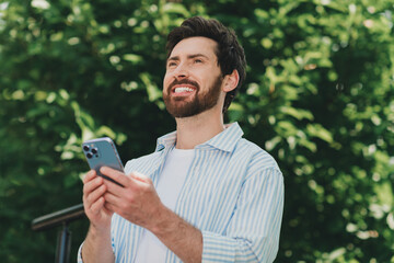 Young man outdoors smiling while using a smartphone, displaying a casual and cheerful look amidst...