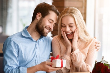 Excited young woman receiving gift from her boyfriend at cozy restaurant. Affectionate millennial couple celebrating Valentine's Day, making surprise for birthday or anniversary