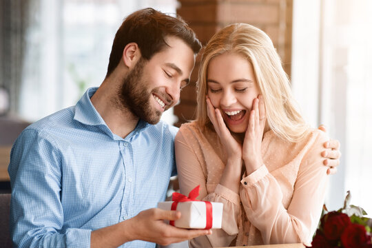Excited young woman receiving gift from her boyfriend at cozy restaurant. Affectionate millennial couple celebrating Valentine's Day, making surprise for birthday or anniversary