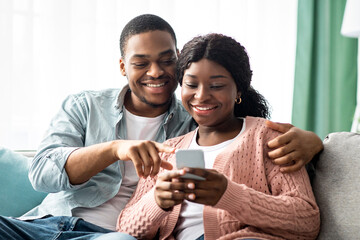 Smiling african american young family using mobile phone together while resting on couch in living room, copy space. Happy black couple checking new shopping application on smartphone