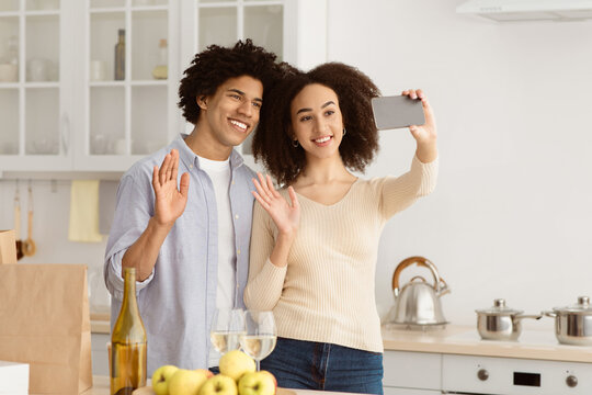 Self-isolation, holiday, anniversary, birthday celebration and video call. Smiling young african american couple make selfie at table with paper bags with food, glasses and bottle of wine in kitchen