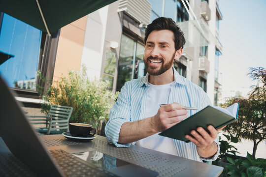 Young man outdoors taking notes in a journal, smiling and surrounded by greenery and sunlight at a street cafe 