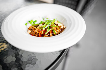 Asian-style noodles with sauce, garnished with green onions, peanuts, and peas, served on a white plate over glass table in a modern dining setting.