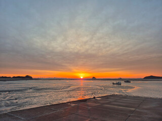 Sunrise over tidal mudflats with glowing orange sky, fishing boats, and small island silhouette on horizon, beautiful coastal seascape nature series