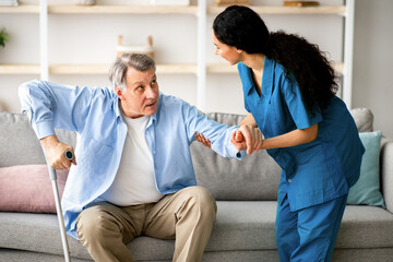 Millennial caregiver helping senior man with crutches to get up from couch at retirement home. Medical worker assisting elderly patient with physical disability, taking care of handicapped male