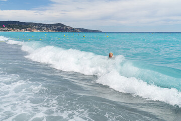 Fototapeta premium A happy, joyful blonde woman plays and swims in the beautiful turquoise waves of the Mediterranean Sea in Nice, France.