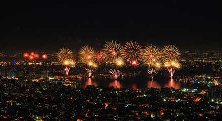 A vibrant firework display illuminates the night sky above a sprawling cityscape and calm body of water.