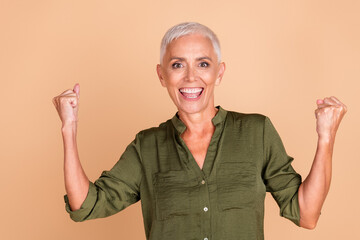 Confident and cheerful senior woman celebrating success with victorious gestures against a beige...