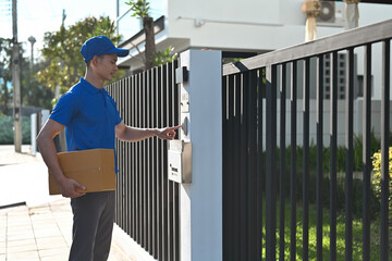 A delivery courier in uniform presses a doorbell while holding a cardboard package outside a gated home