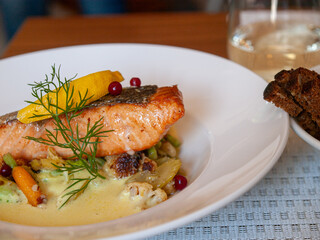 Grilled salmon fillet served with vegetables, creamy sauce and garnish, restaurant dish with wine and bread.