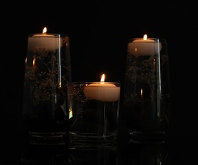 Burning candles and flowers in glasses of water on mirror table against black background