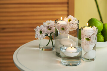Burning candles and flowers in glasses of water on white table indoors