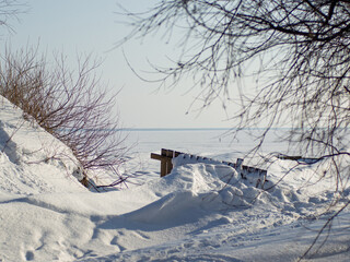 Snow-covered seashore with a wooden bench near frozen sea in winter, peaceful cold season scenery.