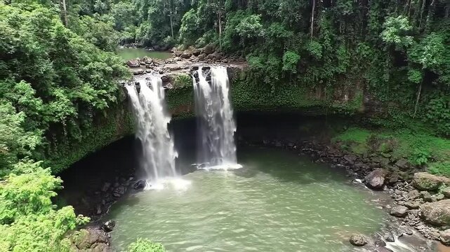 Aerial video footage of a lush green waterfall cascading into a tranquil pool, surrounded by dense tropical rainforest; showcasing vibrant emerald tones and a serene