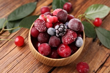 Mix of ripe frozen berries in bowl and green leaves on wooden table, closeup