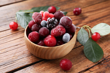 Mix of ripe frozen berries in bowl and green leaves on wooden table, closeup
