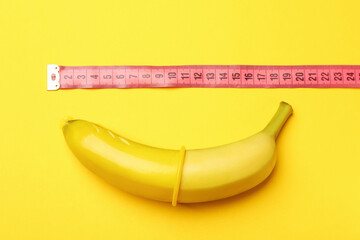 Sex education. Banana with condom and measuring tape on yellow background, top view