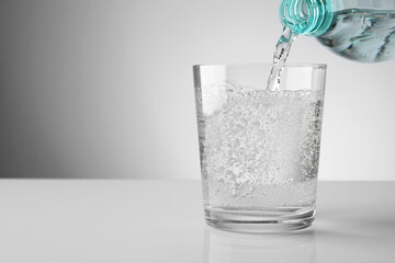 Pouring refreshing soda water from bottle into glass at white mirror table against light grey gradient background, closeup. Space for text