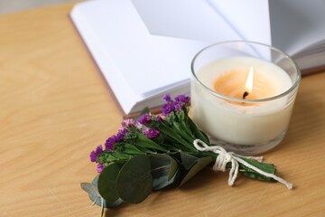 Burning candle, open book and flowers on wooden table indoors, closeup. Space for text