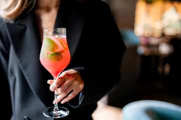 Woman in black blazer holding a glass of pink cocktail with mint garnish in a blurred indoor setting, possibly a bar or restaurant.