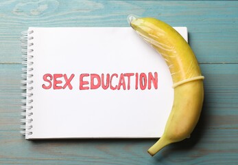 Phrase Sex Education written in notebook and banana with condom on blue wooden table, top view