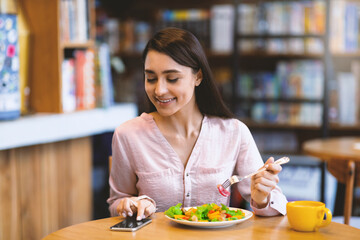 Happy young arab woman texting on smartphone while eating salad in cafe, using an application to send an sms message in her cellphone device. Modern lifestyle concept