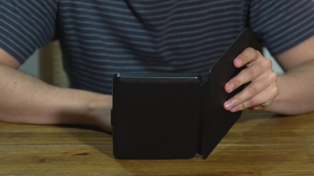 A person in a striped shirt reading an e-book on a black e-reader with a protective case, seated at a wooden table