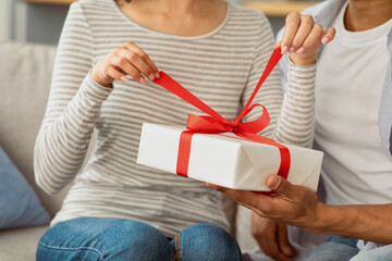 Holiday, anniversary, new year and birthday, celebration together. Millennial african american wife opens gift from her husband, sits on couch in living room interior, cropped, close up, copy space