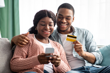 Happy black wife and husband sitting together on couch in living room, holding smartphone and credit card, shopping online, closeup. African american smiling couple using mobile phone at home