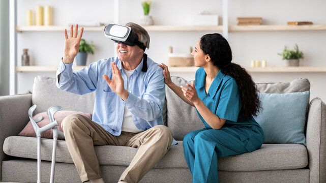 Disabled senior man using VR headset with young nurse, exploring augmented reality at home. Elderly male patient with doctor playing virtual game, having fun with futuristic technologies