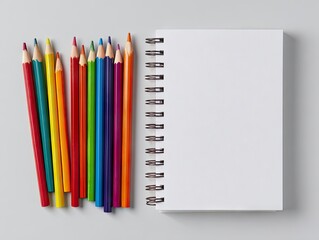 Fototapeta premium A spiral-bound blank notebook rests beside a vibrant collection of colored pencils on a light gray surface