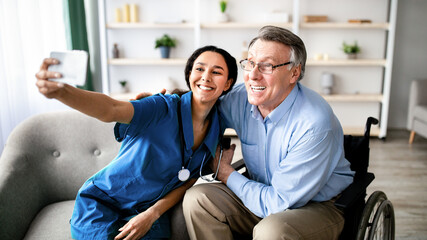 Cheerful impaired senior man in wheelchair taking selfie with young female doctor at retirement home. Disabled older patient making photo with nurse, having fun together indoors