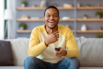 Laughing black guy watching comedy on TV at home, sitting on sofa in living room, holding TV remote. Cheerful african american young man watching something funny show or movie on television