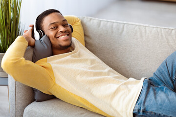 Happy black guy in wireless headset reclining on sofa and smiling while listening to music at home....
