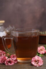 Tasty rose tea in glass cup and flowers on wooden table against brown background, closeup