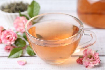 Tasty rose tea in glass cup and flowers on white wooden table, closeup