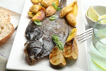 Tasty roasted flounder fish served with potatoes, basil and lemon slices on white marble table, closeup. Homemade seafood dish