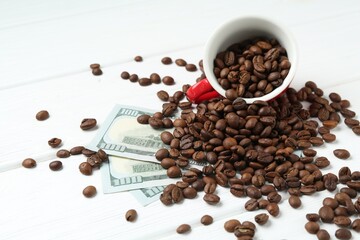 Roasted coffee beans, cup and dollar banknotes on white wooden table, closeup